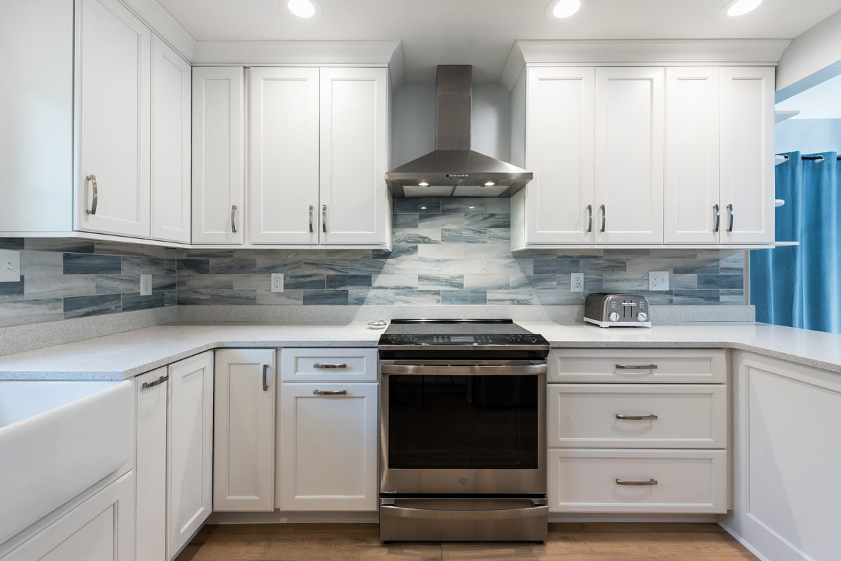 A newly renovated, modern kitchen featuring white shaker cabinets, light wood flooring, stainless steel appliances including a refrigerator and range hood, an undermount farmhouse sink, and a gray and blue subway tile backsplash. A small island with a white countertop is in the foreground.